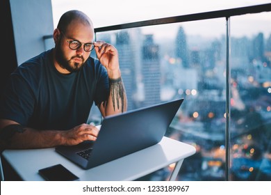 Young handsome skilled IT developer correcting trendy eyeglasses while watching training webinar in webpage on laptop computer.Bearded hipster guy making researches in network sitting in office