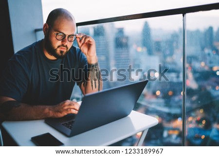 Young handsome skilled IT developer correcting trendy eyeglasses while watching training webinar in webpage on laptop computer.Bearded hipster guy making researches in network sitting in office