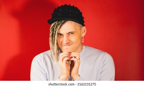 Young handsome sly man with blonde dreadlocks in black cap looking at camera, pursing lips, tapping with folded fingers. Stylish guy with crafty facial expression having idea, plan on red background