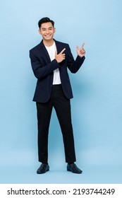 Young handsome smiling Asian man wearing semi-formal suit looking at camera and pointing hands up in blue color studio isolated background