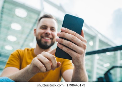 Young handsome smiling bearded man sitting at stadium steps and making bets online on his smartphone using mobile bookmaker application. Sport, gambling, bet, money win concept. - Powered by Shutterstock