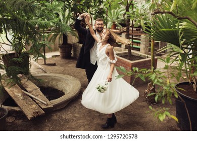 Young Handsome Smiling Groom Happily Dancing With Beautiful Bride In White Dress Holding Little Bouquet Of Flowers In Hand On Their Wedding Spending Time Together In Wonderful Garden Of Old Greenhouse