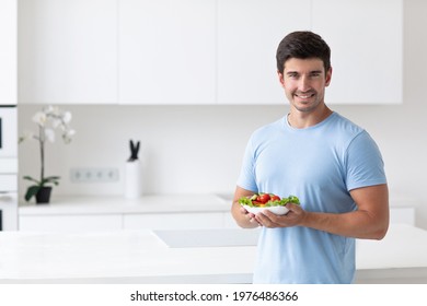Young handsome smiling man in the kitchen with a vegetable salad in his hands, looks good wearing a blue T-shirt. Healthy and vegetarian food concept.