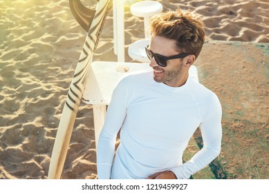 Young Handsome Smiling Man Is Sitting On A Beach Next To His Surfboard Wearing Dark Sunglasses And White Blank Long-sleeve Lycra Shirt. Vertical Mock-up Style.