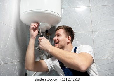 Young Handsome Smiling Man Worker In Uniform Repairing Water Heater Using Screwdriver At Home In The Toilette Professional Repair Service