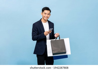 Young Handsome South East Asian Man Holding Mobile Phone And Shopping Bags In Light Blue Studio Isolated Background