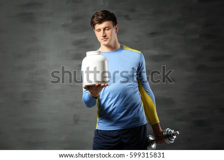 Young handsome sportsman holding bottle of protein and dumbbell on color background