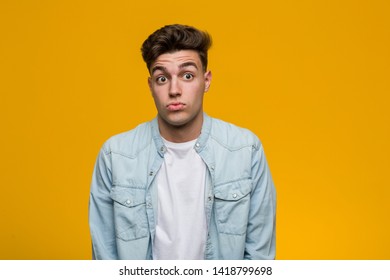 Young Handsome Student Wearing A Denim Shirt Shrugs Shoulders And Open Eyes Confused.