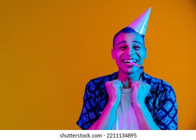 Young handsome stylish enthusiastic boy in birthday cap with birthday pipe in his mouth with raised fists looking at camera , while standing under blue and pink light over isolated orange background - Shutterstock ID 2211134895