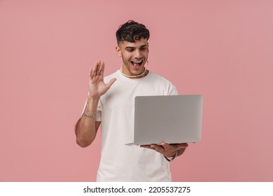 Young Handsome Stylish Enthusiastic Man Holding Laptop Looking And Waving To It , While Standing Over Isolated Pink Background