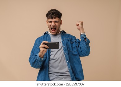 Young handsome stylish enthusiastic man holding phone doing winner gesture with raised fist and opened mouth , while standing over isolated brown background - Shutterstock ID 2205537307