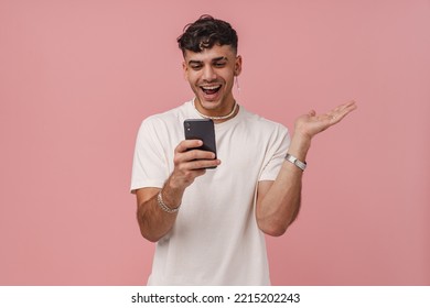 Young Handsome Stylish Enthusiastic Man With Opened Mouth And Raised Hand Holding Phone And Looking At It , While Standing Over Isolated Pink Background