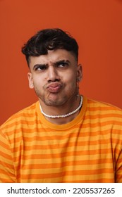 Young Handsome Stylish Frowning Man With Puckered Lips And Piercing In Necklace, Standing Over Isolated Orange Background