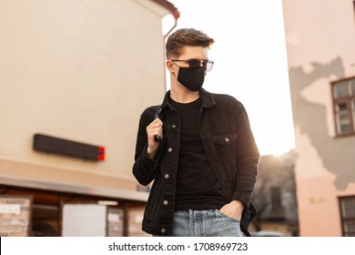 Young Handsome Stylish Man Model In Stylish Black Denim Jacket With Backpack In Vintage Sunglasses In Black Trendy Protective Mask Poses In City On Bright Spring Sunny Day. Cool Guy Hipster In Street.