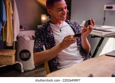 Young Handsome Stylish Smiling Boy Looking At Polaroid Pictures, While Sitting By Table In Cozy Room At Home