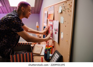 Young Handsome Stylish Smiling Boy Pinning Picture On Cork Board, While Standing In Cozy Room At Home