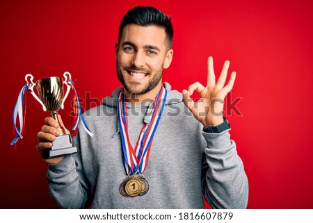 Young handsome succesful man holding trophy wearing medals over red background doing ok sign with fingers, excellent symbol
