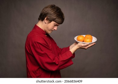 Young Handsome Tall Slim White Man With Brown Hair Humbly Offering Oranges In Red Shirt On Grey Background