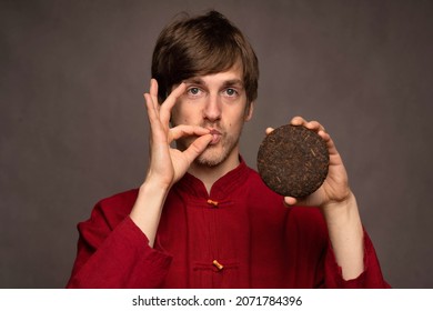 Young Handsome Tall Slim White Man With Brown Hair Doing Chefs Kiss Sign Presenting Pu Erh Tea Cake In Red Shirt On Grey Background