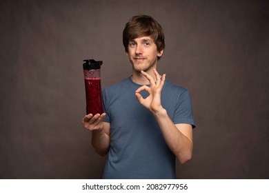 Young Handsome Tall Slim White Man With Brown Hair Holding Smoothie Bottle Making Ok Sign In Grey Shirt On Grey Background