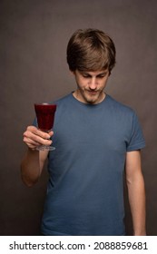 Young handsome tall slim white man with brown hair holding wine cup facing down in grey shirt on grey background