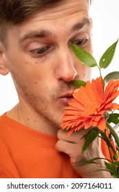 Young Handsome Tall Slim White Man With Brown Hair Orange Flower Really Close To His Face With Orange Shirt Isolated On White Background