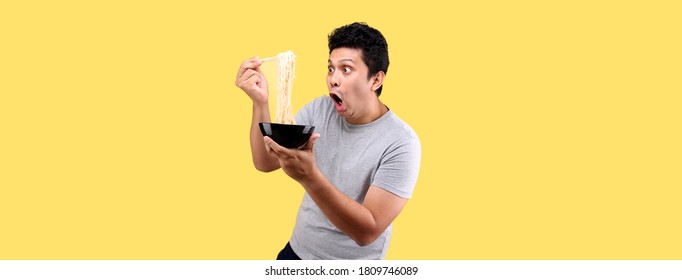 Young Handsome Trendy Asian Man Eating Yummy Hot And Spicy Instant Noodle Using Chopsticks And Bowl Isolated On Yellow Background In Studio With Copy Space.