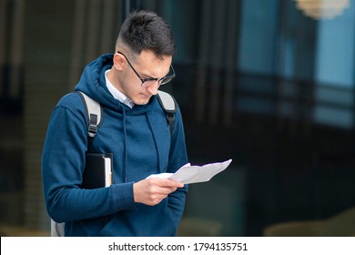 Young Handsome Upset Dissatisfied Guy, Serious Focused Student With Backpack In Glasses Standing With Book, Reading Papers, Documents, Looking At List With Results Of Text, Exam With Concentrated Look