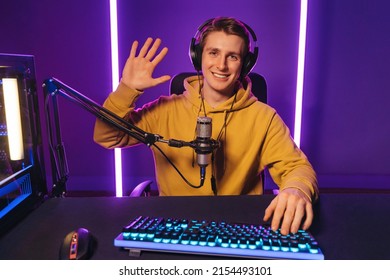 Young handsome vlogger pro gamer waving hand to camera, says hello to his subscribers and followers, smiling on camera. He recording live stream of video game playthrough at home at night. Cyber sport