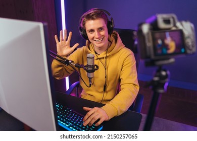 Young handsome vlogger professional gamer waving hand to camera, smiling, greeting his followers with hand gesture while shooting vlog and playing video games on his computer at home. Cyber sport