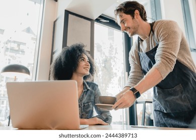 Young handsome waiter servicing black girl customer with laptop in cafe. Customer service area in small business. Female freelancer ordering hot beverage during work break in restaurant - Powered by Shutterstock