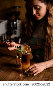 young handsome woman neatly decorates glass with bumble coffee cocktail with slice of dry orange and mint leaf