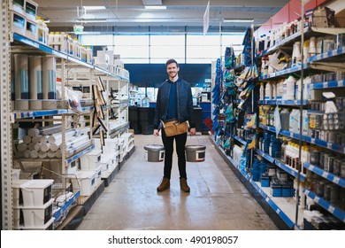 Young Handyman Posing In A Hardware Store Standing Smiling At The Camera In The Aisle Between Racks Of Merchandise