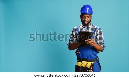 Young handyman using digital tablet on camera, preparing to start constuction contract. Male carpenter in uniform browisng online social media page on modern gadget, building site.