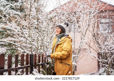 Young happy adult caucasian woman smiling in a hat and yellow jacket enjoying the snowfall on the terrace of a country house in forest