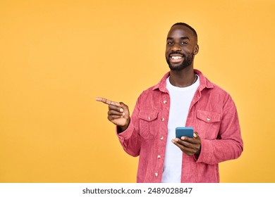 Young happy African American Black hipster man with cellphone in hands standing isolated on yellow background holding smartphone using mobile cell phone pointing aside advertising betting app. - Powered by Shutterstock