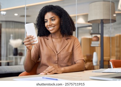 Young happy African American business woman using mobile phone sitting in corporate office. Professional female execuitve or entrepreneur looking at smartphone working with cellphone in hands.