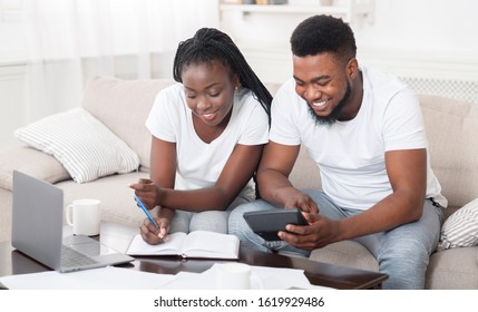 Young Happy African American Couple Planning Family Budget Together, Sitting On Sofa At Home, Using Calculator And Taking Notes