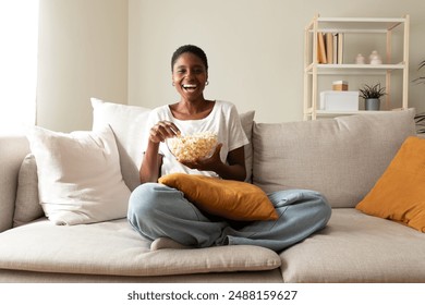 Young happy African American female eating popcorn, sitting on the couch looking at camera laughing. Black woman watching movie at home. Leisure activity. - Powered by Shutterstock