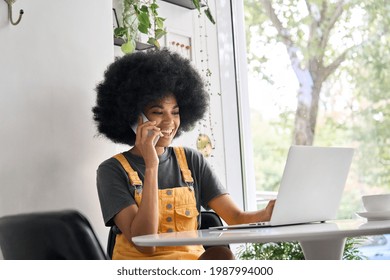 Young Happy African American Generation Z Student With Curly Afro Hair Sitting In Cafe Speaking On Mobile Cell Phone, Using Laptop, Working Learning Remotely Indoors In Home Office.