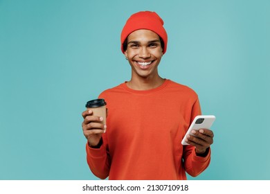Young happy african american man in orange shirt hat hold takeaway delivery craft paper brown cup coffee to go use mobile cell phone isolated on plain pastel light blue background studio portrait.