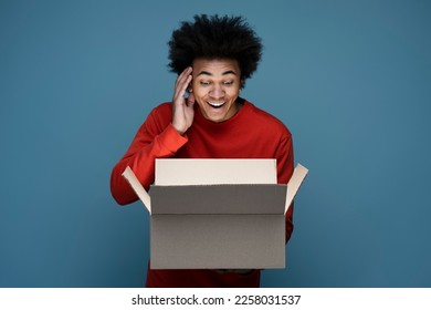 Young happy African American man influencer unpacking delivery box isolated on blue background. Excited customer opening package with birthday gift. Online shopping, sale concept 