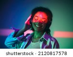 Young happy african american woman in black headphones listening to music, enjoying high quality sound while standing under purple neon glowing light in studio and smiling happily. Musicial equipment