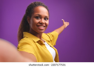Young happy African American woman points back with hand inviting you to come in or calls for visit and films himself on camera and takes selfie standing in purple studio. Headshot screen view - Shutterstock ID 2207177531