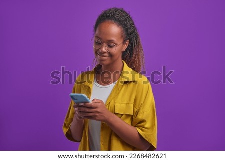 Young happy African American woman uses phone and is touched by reading sentimental story about romantic relationship on social network stands on lilac background. Smartphone, technology, emotions