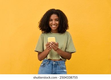 Young happy African gen z teen girl standing at yellow wall looking at camera holding smartphone. Smiling Black woman using mobile phone advertising summer shopping sale or mobile apps. Portrait. - Powered by Shutterstock