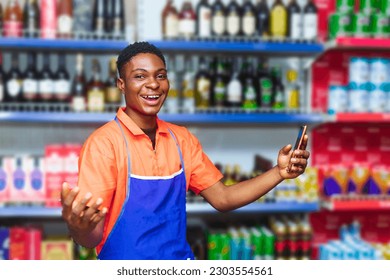 young happy African sales male supermarket attendant on orange attire holding phone while wearing a blue apron - Shutterstock ID 2303554561