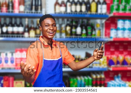 young happy African sales male supermarket attendant on orange attire holding phone while wearing a blue apron
