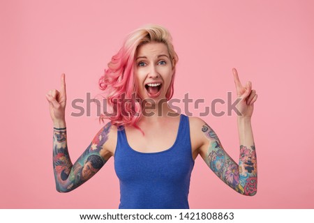 Young happy amazed beautiful pink haired woman in blue t-shirt with arms raised, hears unbelievable news, with wide open mouth in surprise, stands over pink background and pointing up at copy space.