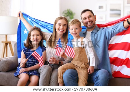 Young happy american family parents and two little kids sitting on sofa at home with flags of united states and smiling at camera while celebrating Independence Day. Patriotic US holiday concept